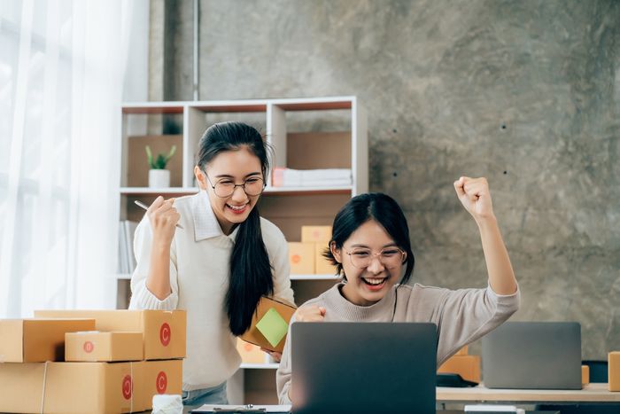 Two e-commerce business women in front of a laptop with boxes of items next to it