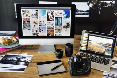 Computers, photos, and a DSLR camera sitting on a wooden desk, symbolizing photo editing