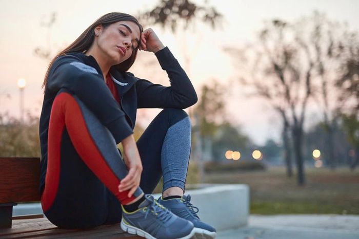 A woman in workout clothes resting on a bench in a park with her eyes closed