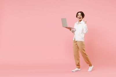 A woman with short brown hair, wearing a white button-down long-sleeve shirt, khaki pants, and white sneakers, standing in a room with a coral pink interior. She is holding her silver laptop in her one hand, with the other arm lifted and the hand balled into a fist. She has a very ecstatic expression on her face. 