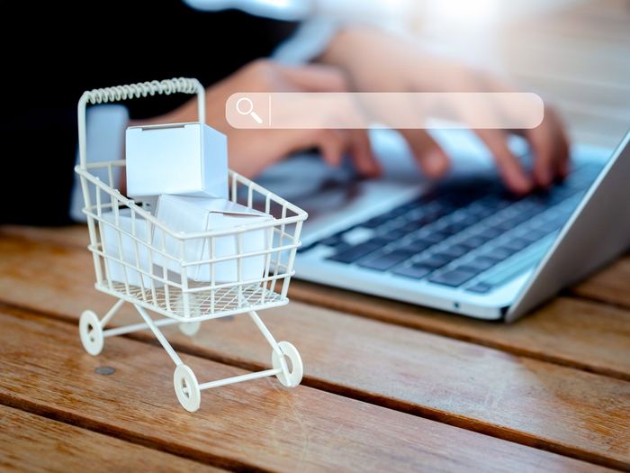 Laptop on a wooden table with a person shopping online and a small white shopping cart on the table