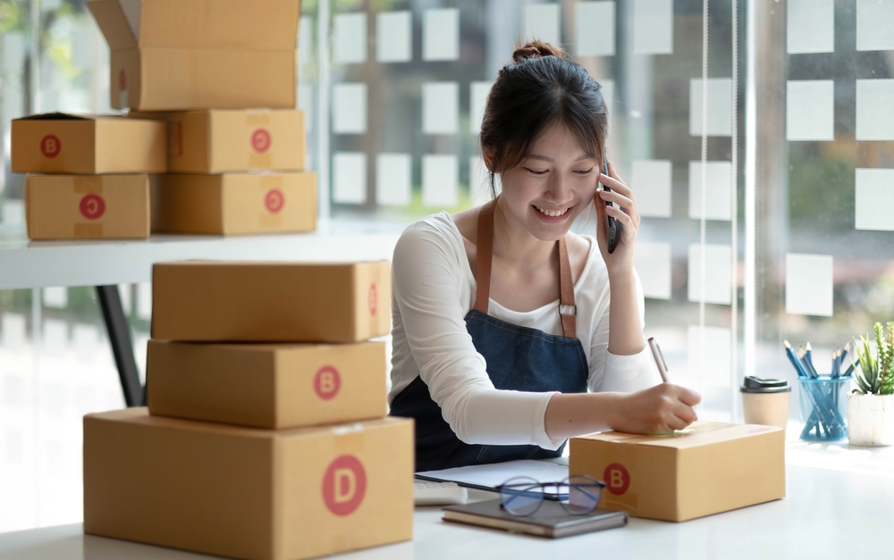 Woman wearing apron and talking on phone and writing information down next to packed boxes