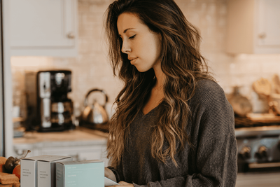 A woman standing in her kitchen looking at a box on ben wa balls and a Kegel device.