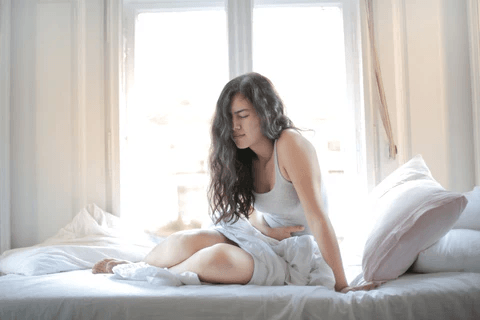 Vaginismus A woman sitting on a bed near a bright window, holding her lower abdomen with an expression of discomfort or pain