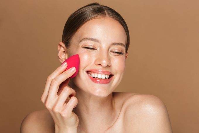 A woman is smiling while holding a pink concealer sponge.