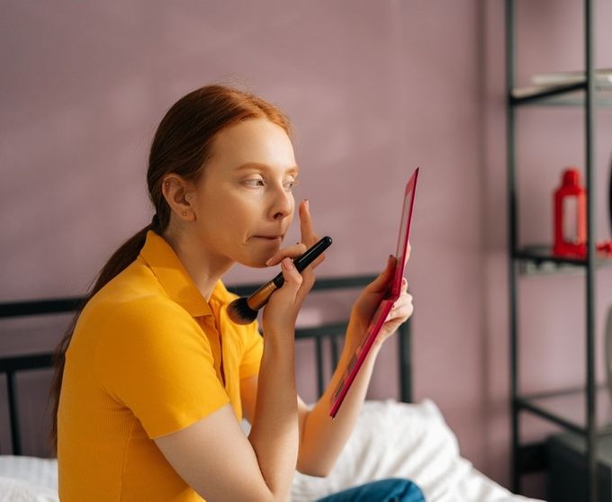 A woman sitting on a bed applying makeup.