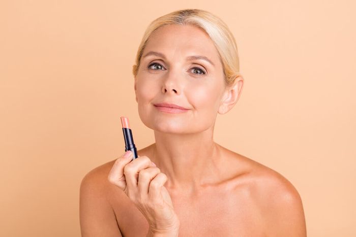 A woman smiling and holding a lipstick in front of her face against a nude-colored background