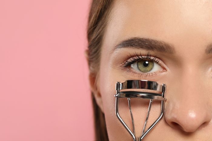 A woman holding an eyelash curler to half her face showing with a pink background
