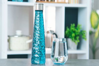 A glass water bottle standing next to a full glass of water, with kitchen items visible in the background.