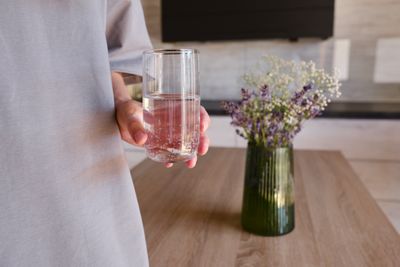 Glimpse of a torso, with the person wearing a grey shirt and holding glass of water, with a green vase populated with dainty purple and white flowers on a wooden table in the middle of the room