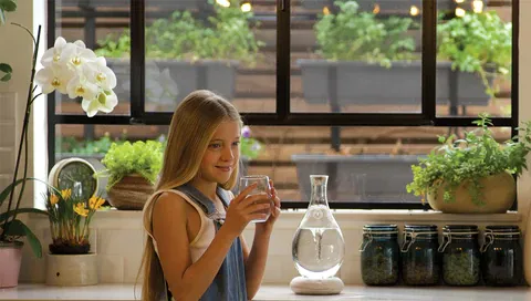Young girl holding a glass of water in a kitchen, water jar on the countertop, pot plants on window seal