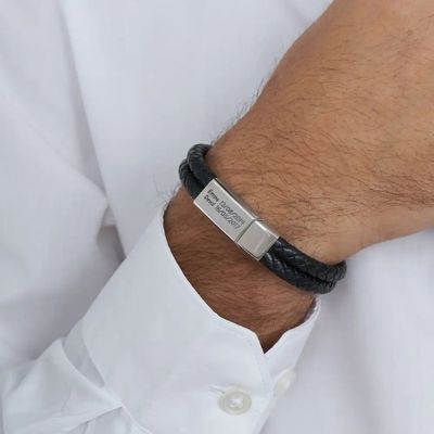 A black woven leather bracelet with a metal bar inscribed with names and dates on a man's wrist