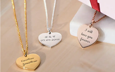 Three necklaces with inscribed heart-shaped pendants in gold, silver, and rose gold on a wooden table