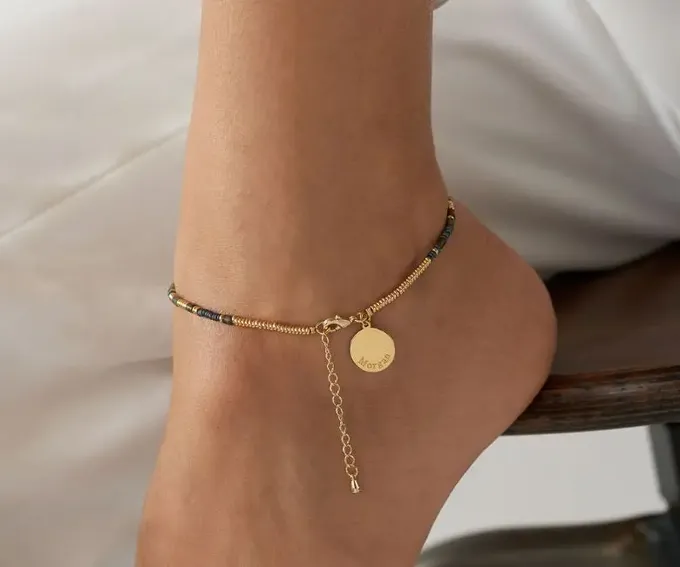 a woman wearing a gold ankle bracelet with a disc charm