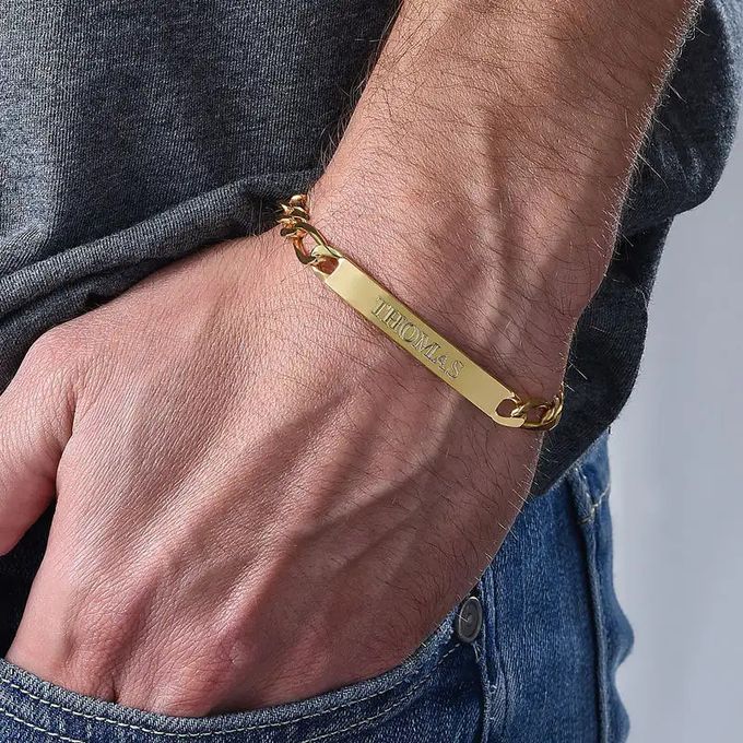 Close up of a man holding his hand in his pocket while wearing a gold name bracelet with a plate that reads "Thomas" around his wrist