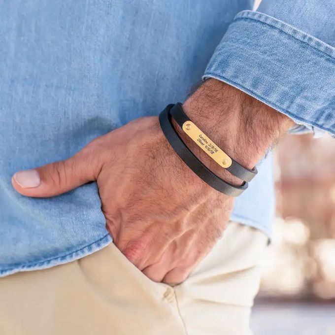 A black leather bracelet with gold plating inscribed with names and dates around a man's wrist