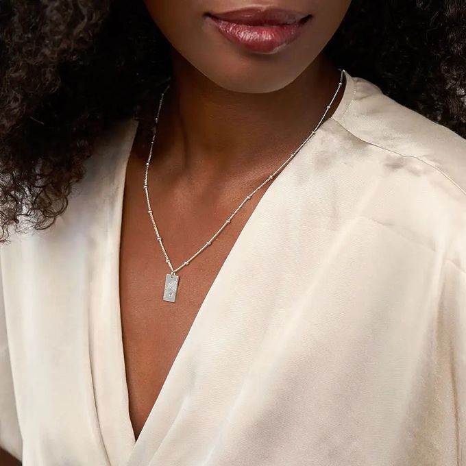 Woman wearing a silver necklace with a rectangular pendant inscribed with a name and embellished with a birthstone and flower