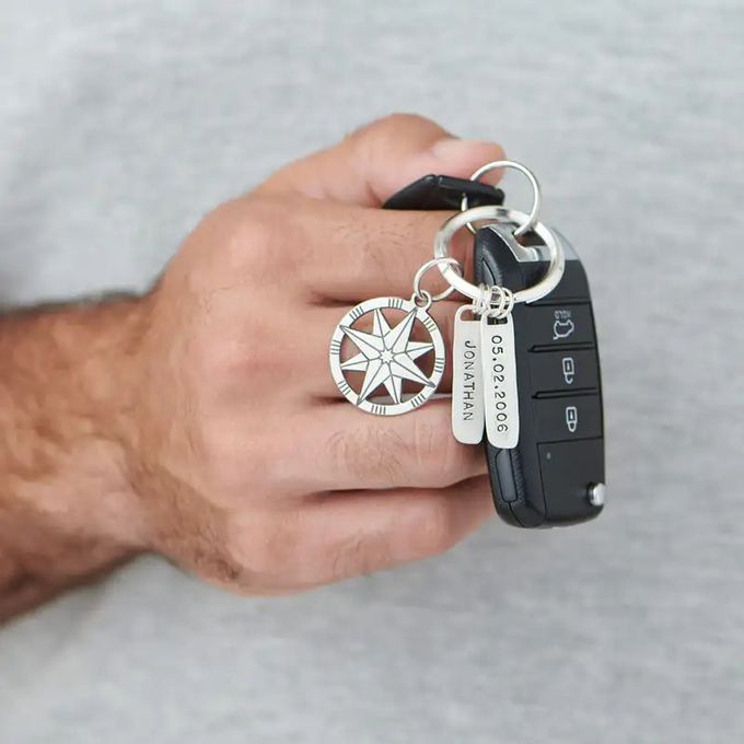 Close-up of a man holding his car keys with a silver key chain with a compass pendant and name-inscribed bars