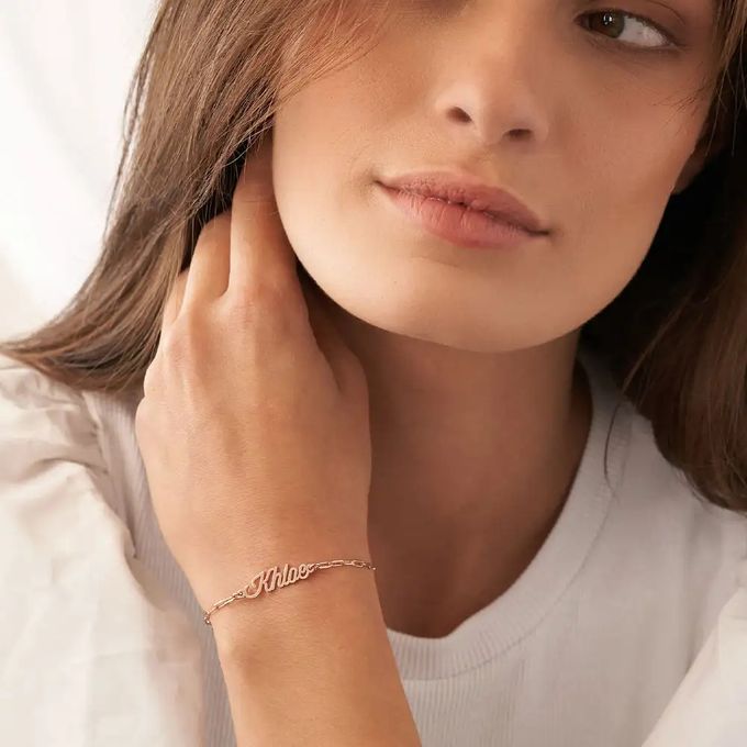 A paperclip bracelet in rose gold with a nameplate that reads "Khloe" in a cursive font around a woman's wrist