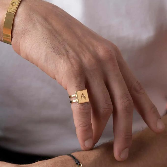 Close-up of a man's hand wearing a gold double-band rectangular ring inscribed with "V"