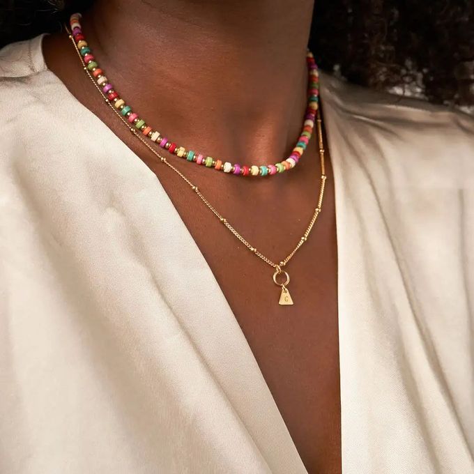 Woman wearing a layered necklace with colorful beads, gold plating, and a triangle-shaped pendant with an initial engraved