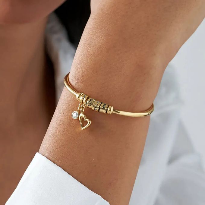 An open bangle bracelet with engraved beads and a heart charm and crystal around a woman's wrist