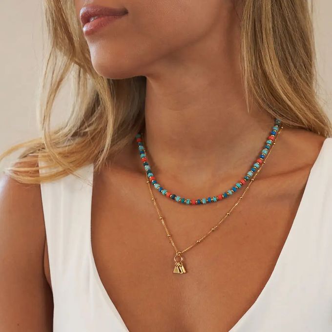 Blonde woman wearing a layered gold chain and colorful beads necklace with initial triangle pendants