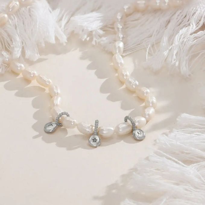 A pearl necklace with silver initials on a white background surrounded by white feathery fabric