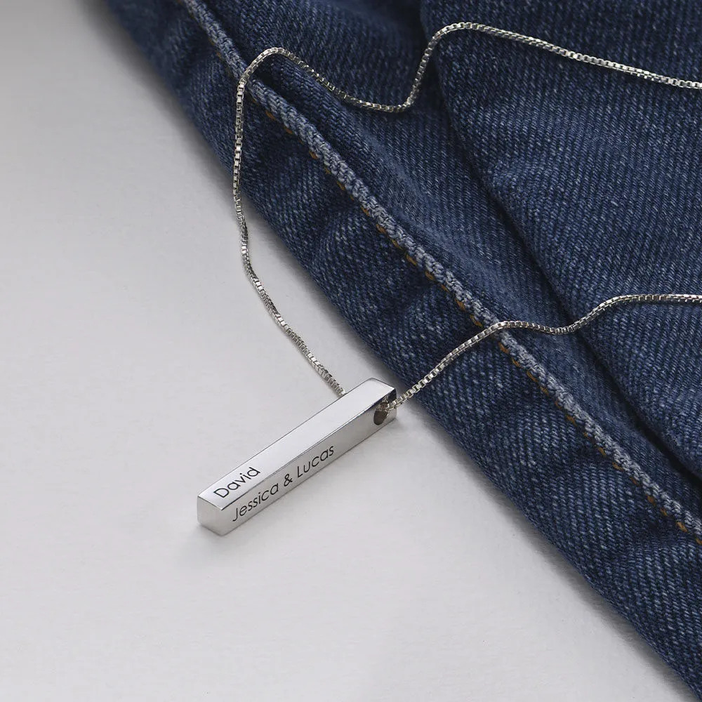 Silver necklace with a vertical bar pendant inscribed on two sides displayed over a jeans material