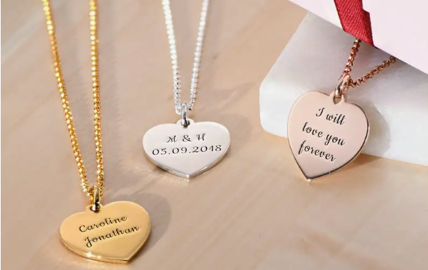 Three necklaces with inscribed heart-shaped pendants in gold, silver, and rose gold on a wooden table