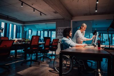 Two professionals working together at a desk in a coworking space, the woman standing and the man seated in his wheelchair.