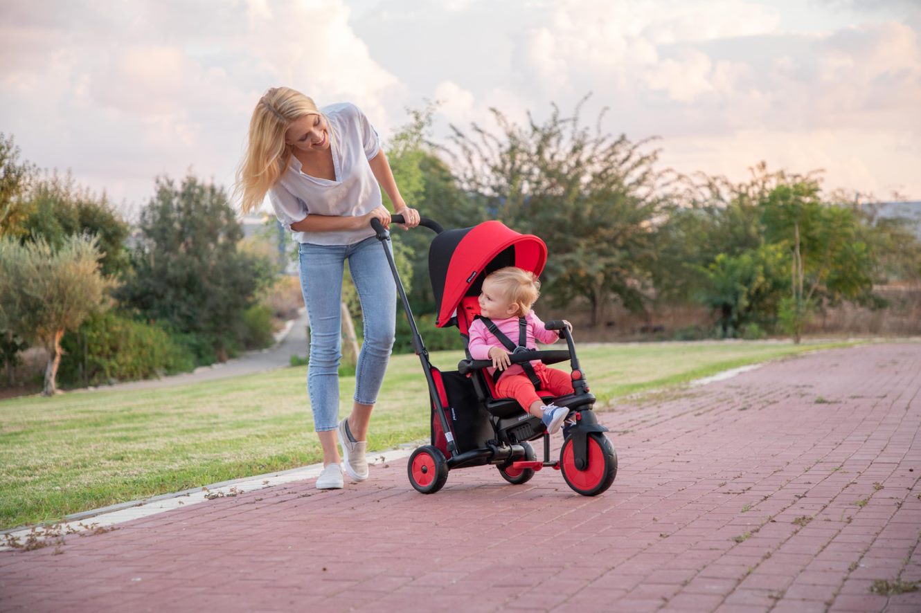 Woman walking a baby in a red stroller trike on a path in a park