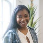 a smiling woman in a denim jacket stands in front of a potted plant