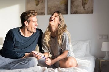a man and a woman over 35 sitting on a bed smiling and waiting for pregnancy test results