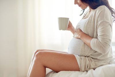 a pregnant woman sitting on a bed holding a coffee cup