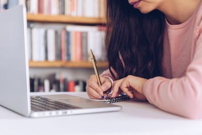 a woman is writing on a notepad in front of a laptop