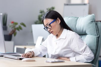 a woman sitting at a desk working on a computer, experiencing period pain at workplace