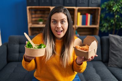 A woman holds a bowl of salad and  a box of fried food.
