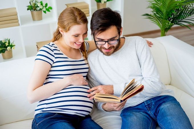 a pregnant couple sitting on a couch reading a book