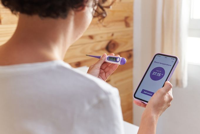 a woman holding a smart phone and a BBT thermometer measuring her BBT and monitoring her OPK results