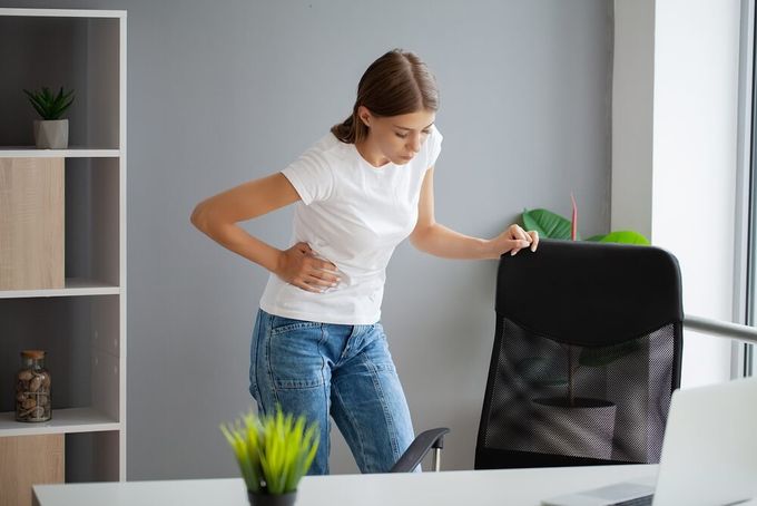 a woman holding her stomach while standing in an office, experiencing strong period pain and cramps
