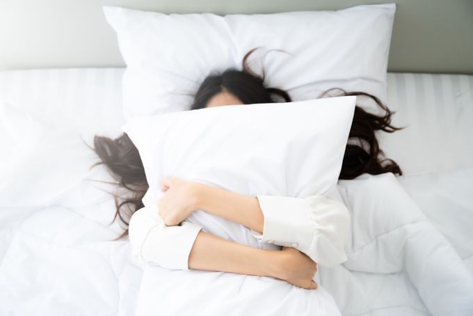 A woman laying in bed under a white pillow feeling moody and emotional during ovulation.
