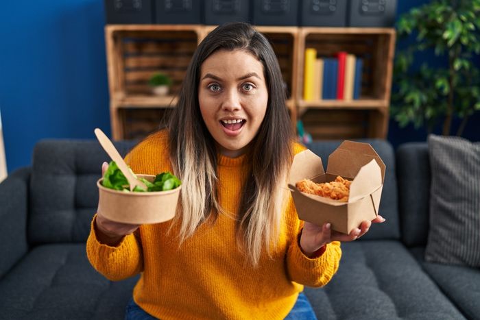 A woman holds a bowl of salad and  a box of fried food.