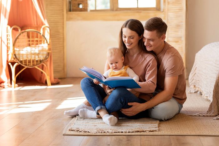 A man and woman are sitting on the floor reading a book to a baby.