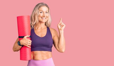 Woman in exercise clothes holding a yoga mat and smiling while pointing up with her finger