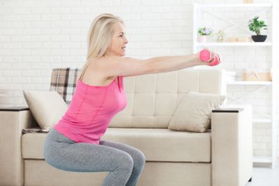 Blonde middle-aged woman in exercise attire doing squats in her living room holding small dumbbells