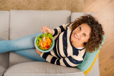 Top view of a woman sitting on a couch, smiling, and eating a salad
