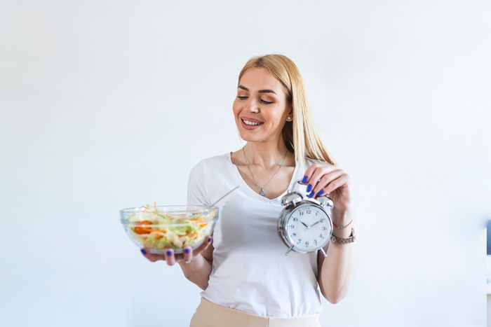 Blonde woman holding a bowl of salad in one hand and a clock in the other and smiling while looking at the salad