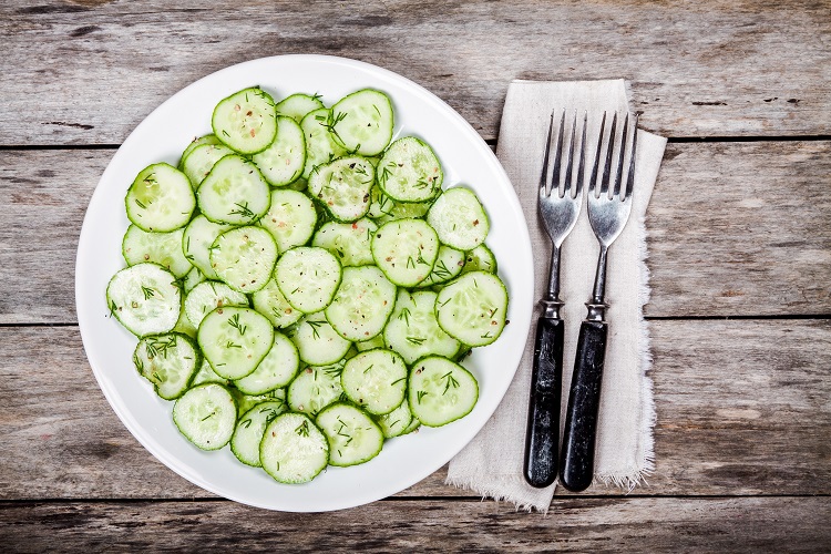 Top view of a plate with cucumber salad next to utensils on a wooden table