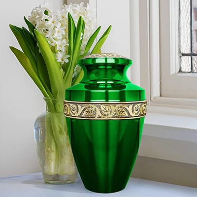 An extra large green cremation urn on display near a windowsill, with  a vase of flowers placed behind it.